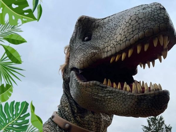 Close-up of a realistic dinosaur puppet with sharp teeth and textured scales, set against a cloudy sky with green jungle leaves framing the corner.