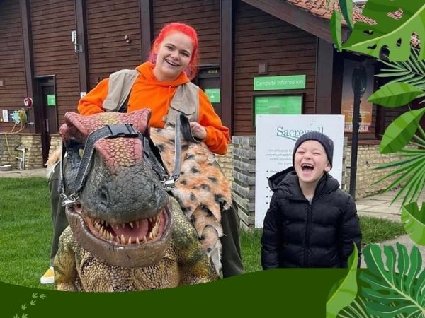 Performer with bright red hair posing with a realistic dinosaur puppet and a laughing child outside a building at Sacrewell, surrounded by playful jungle-themed graphics.